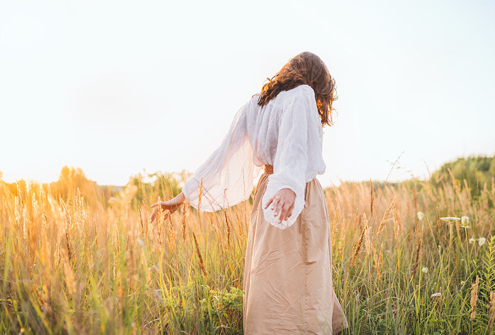 A woman in a white shirt and beige skirt standing in a field with sunlight filtering through.