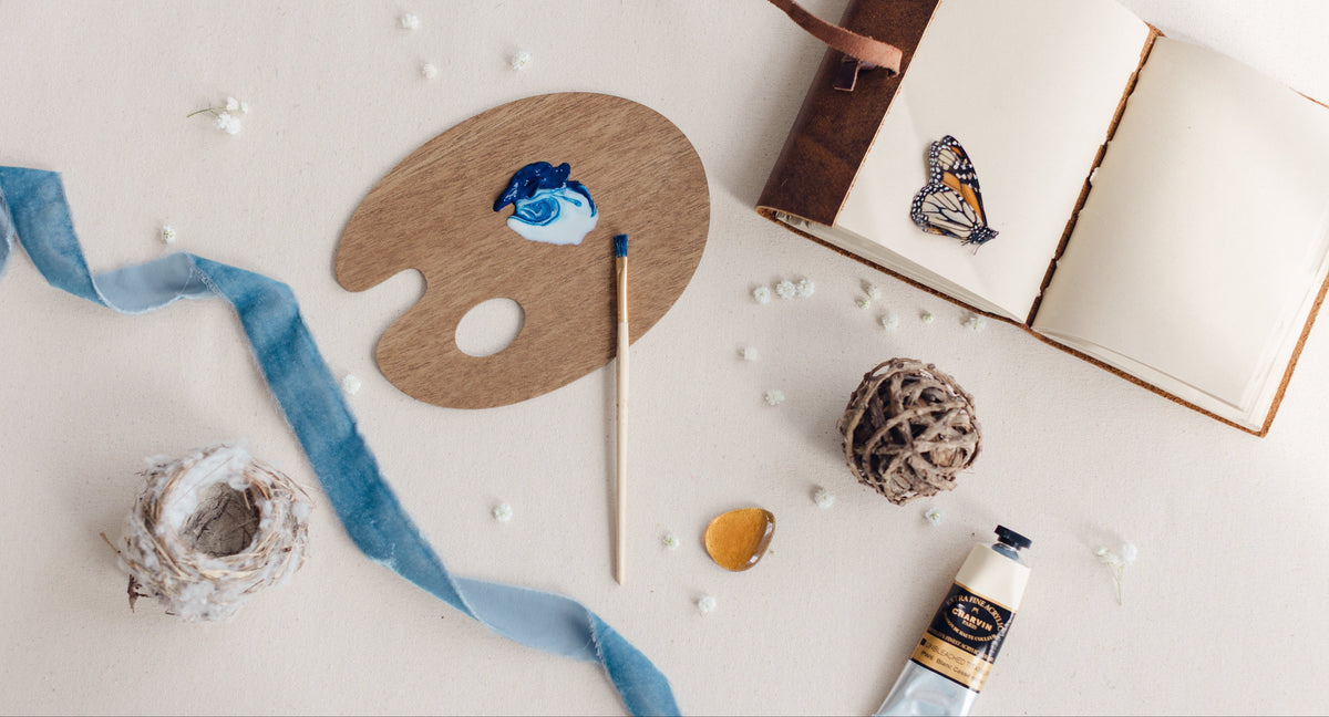 A styled flat-lay of painting and art supplies on a light cream background, including a wooden artist palette with blue paint, a paintbrush, a tube of white paint, a blue ribbon, a small ball of thread, a rolled piece of paper, and an open book with a butterfly illustration from artist Nicole Z Edwards. 