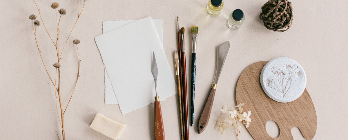 A styled flat-lay of painting and art supplies on a light beige background, including a wooden palette with a white painted sample, paintbrushes, small bottles, dried flowers, paper, and a palette knife from artist Nicole Z Edwards. 