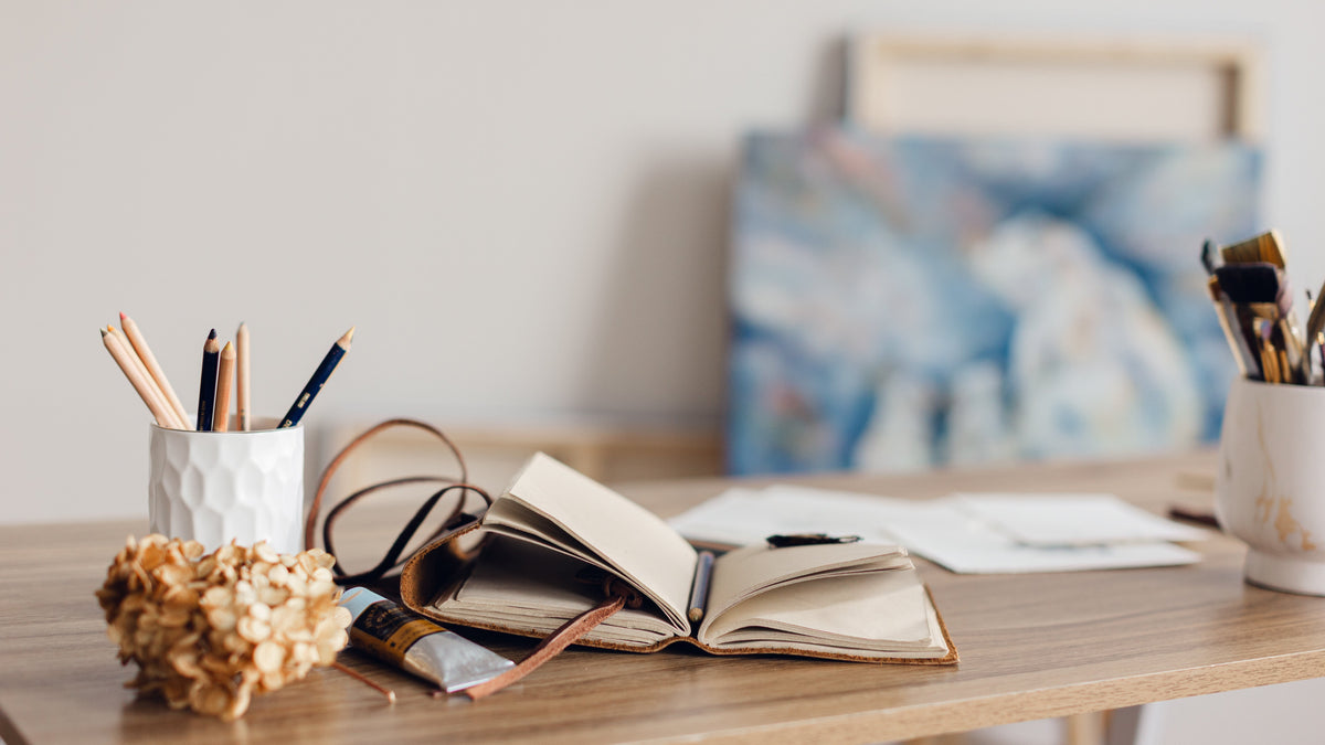 An open notebook with a brown leather bookmark lies on a wooden desk, surrounded by a white pencil cup, eyeglasses, a small dried flower arrangement, and another cup with paintbrushes, with a blurred painting in the background from artist Nicole Z Edwards. 