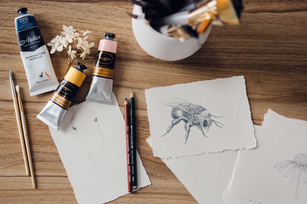 Drawings by mixed media artist Nicole Z Edwards of drawings of a bee, horse portrait, and flower with a vase, paints, paintbrushes and pencils on a wooden table. 