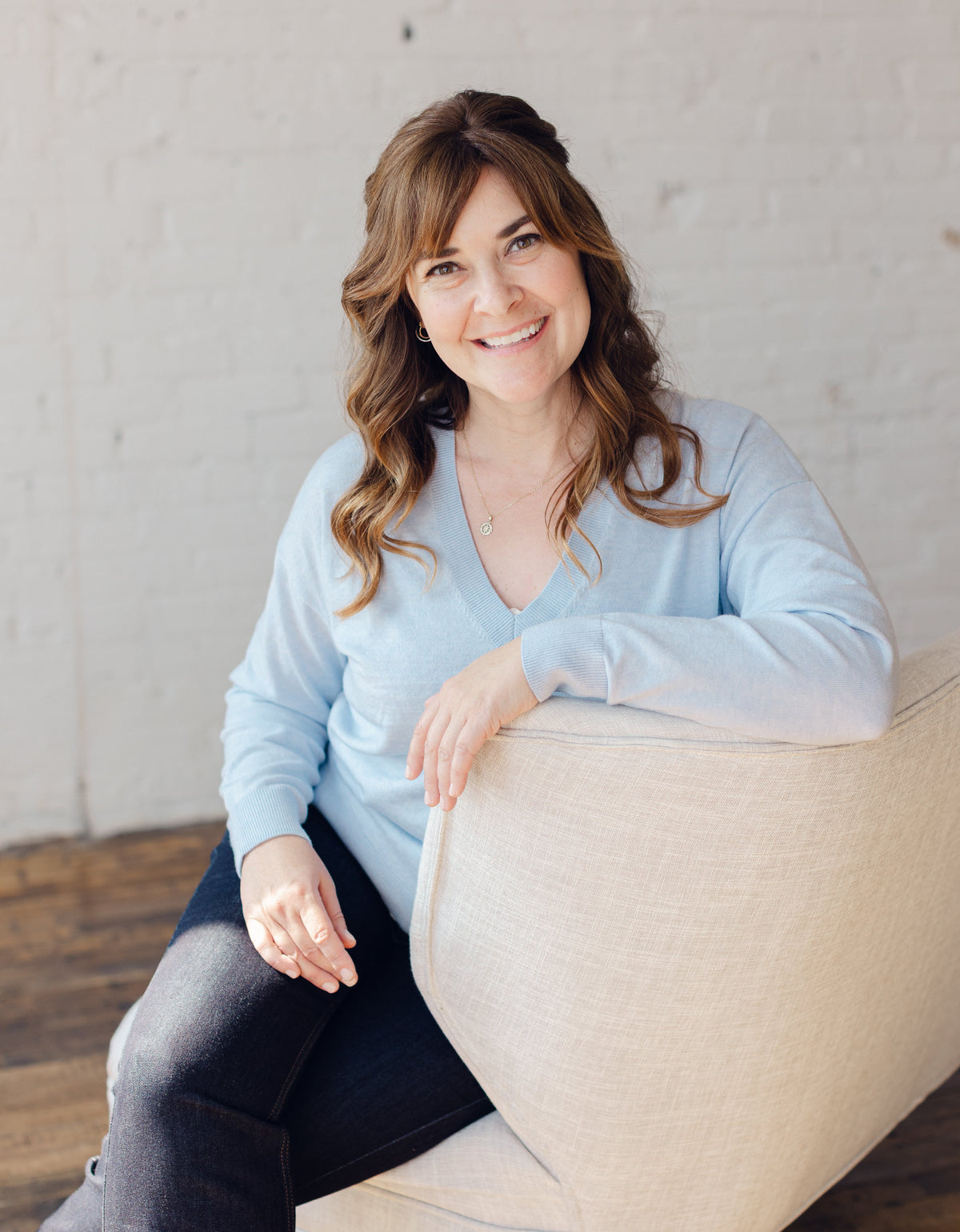 Nicole Z Edwards, a smiling woman with wavy brown hair sits on a beige upholstered chair in a bright indoor setting with a white brick wall and wood floor. She is wearing a light blue long-sleeve V-neck top and dark pants.