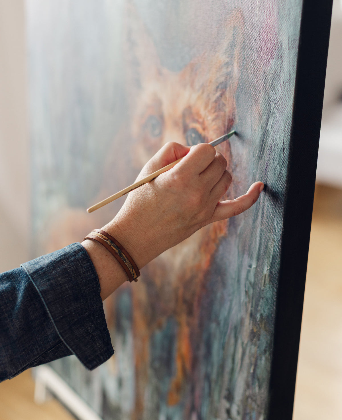 A close-up of mixed media artist Nicole Z Edwards' hand holding a paintbrush and painting on a large canvas of a fox shown in a softly lit indoor setting.