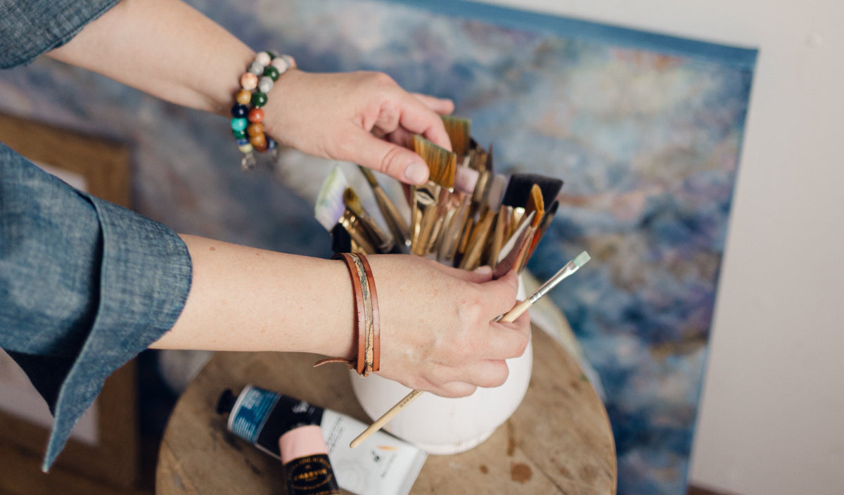 Mixed media artist, Nicole Z Edwards, in a denim shirt selects paintbrushes from a white vase sitting on a rustic wooden stool, with a blue blurred painting leaning against a white wall in the background.