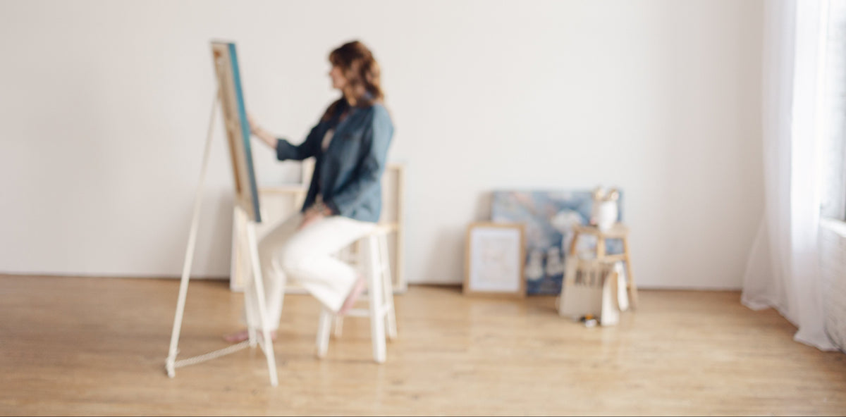 Mixed media artist, Nicole Z Edwards, sits on a white chair at an easel, painting on a canvas in a bright, minimalist art studio with light wood floors and white walls.