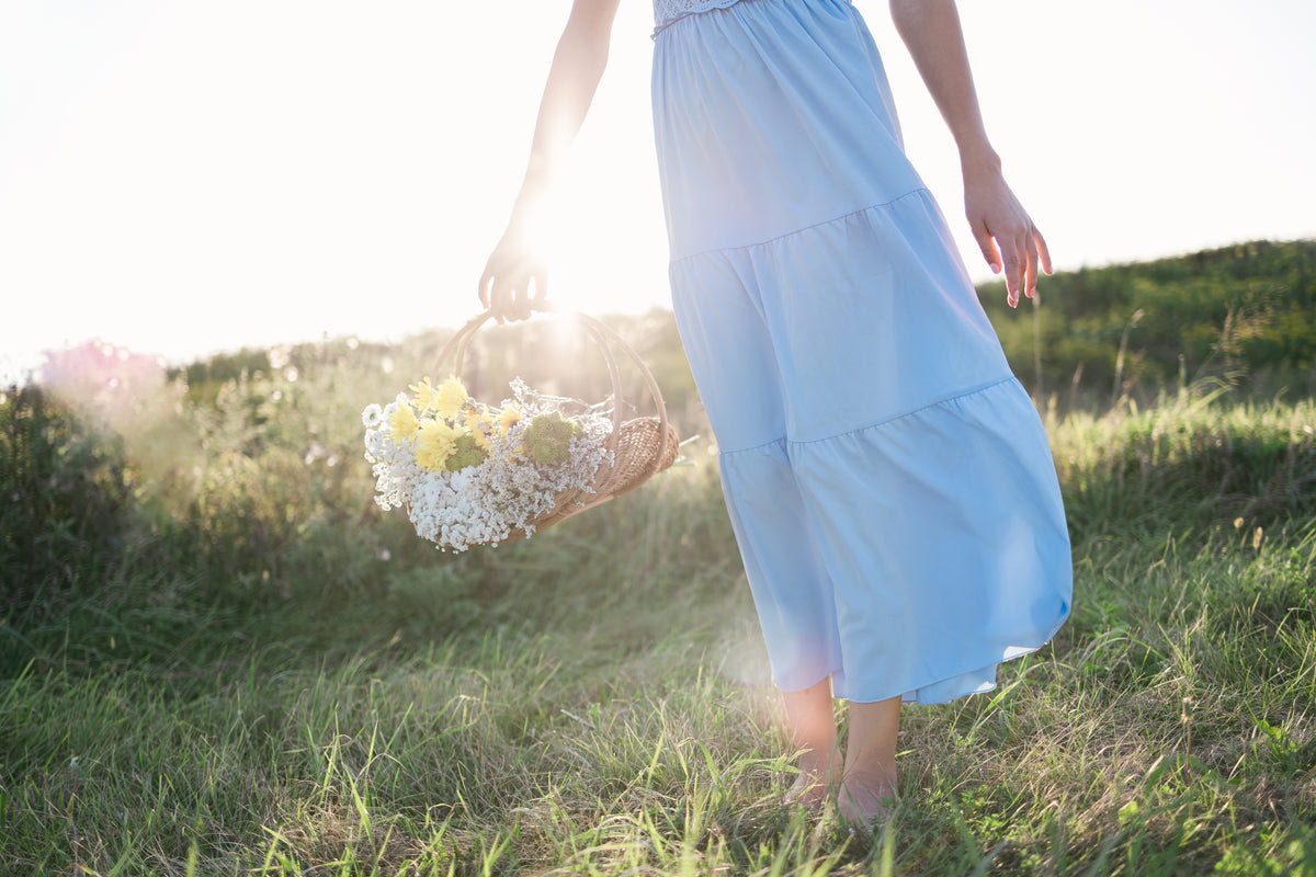 A woman in a light blue tiered maxi dress walking barefoot in a grassy field, holding a woven basket filled with white and yellow wildflowers. The image is shot from behind with bright backlighting and a soft-focus natural background.