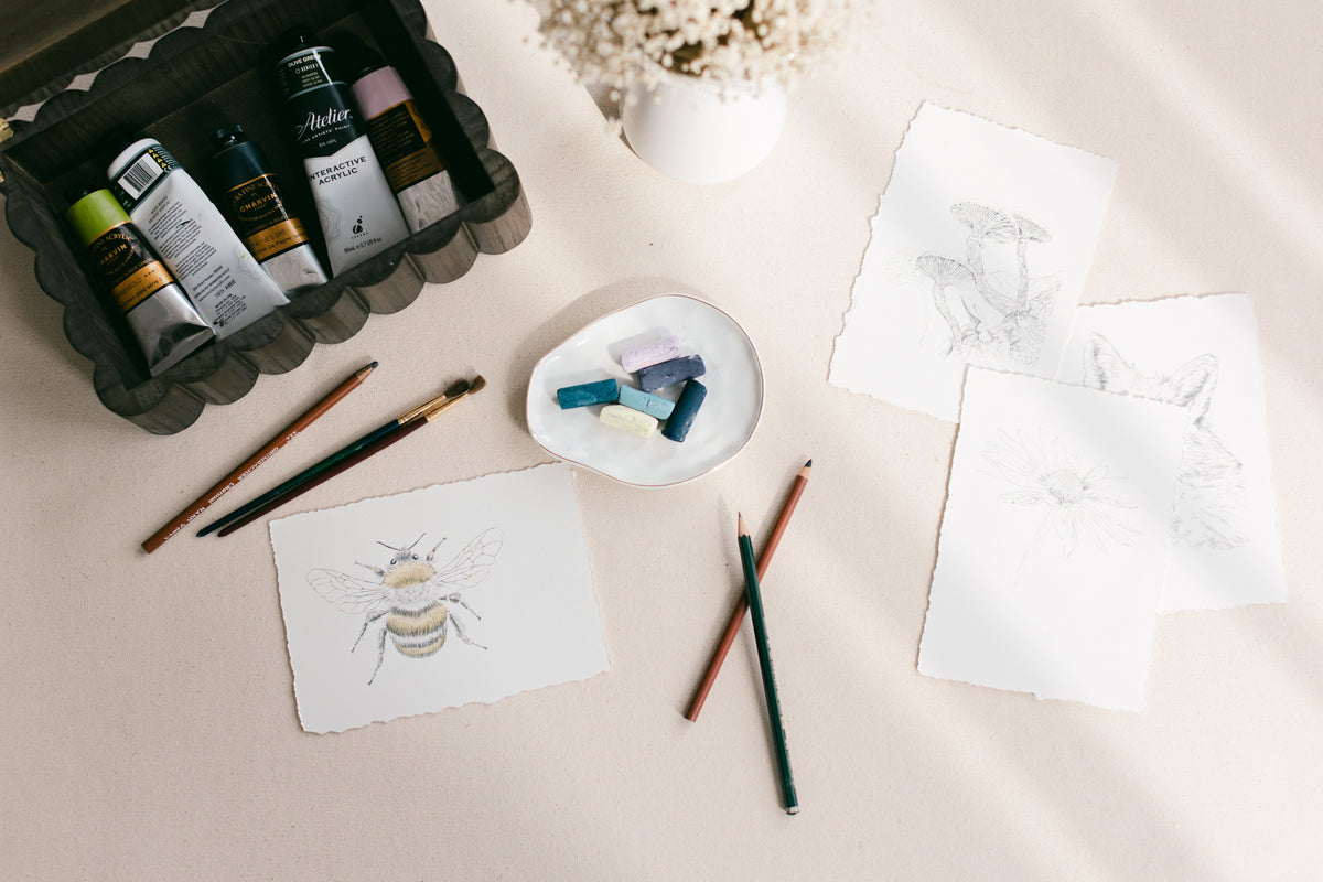 A styled flat-lay of mixed media artist, Nicole Z Edwards featuring a bee sketch and others with pastels, paints, and pencils on beige background.