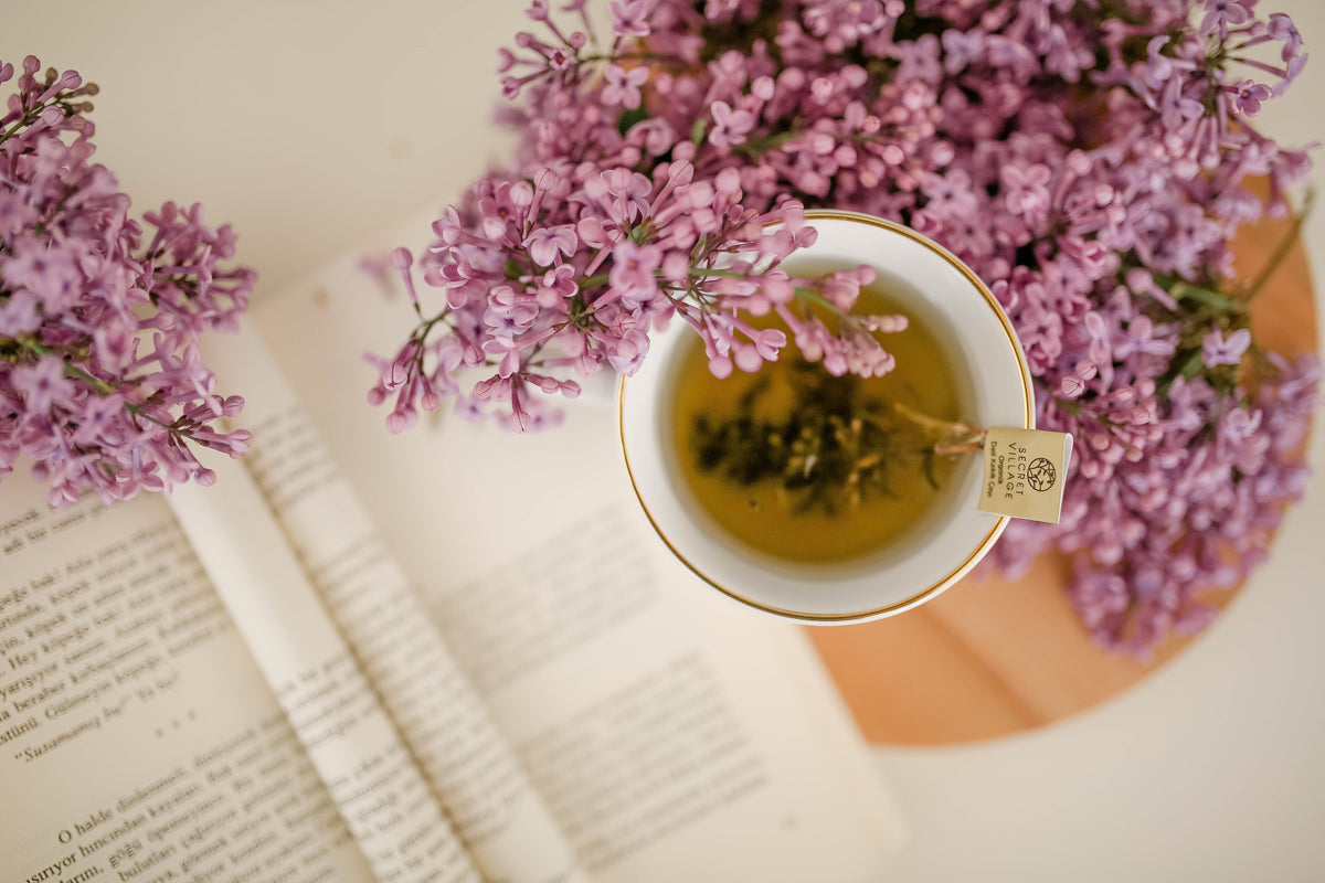 A top-down view of a white teacup with gold trim filled with tea, placed on an open book and surrounded by purple flowers. A tea bag tag is visible in the cup.